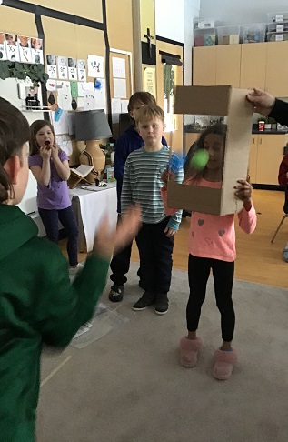 5 School age children - one holding a cardboard box with a window cutout and protective transparent film in the window. One school age child throwing a ball at the window while 3 other school age children are standing and observing
