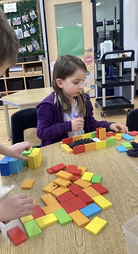 2 school age children sitting at t table with multiple small blocks with various colours stacking and building