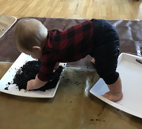 An infant reaching into a tray filled with mud