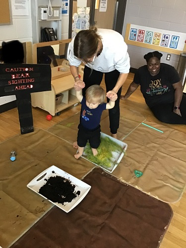 An educator helping a child set into a tray filled with leaves and grass
