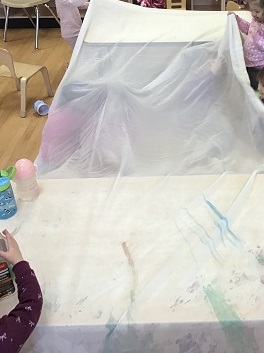 Toddler children sitting under a thin fabric that is raised above the children by being placed on two tables