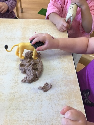 3 Toddler children sitting at the table with brown playdough, one child placing the lion figure into the brown playdough with some circular prints on parts of the playdough