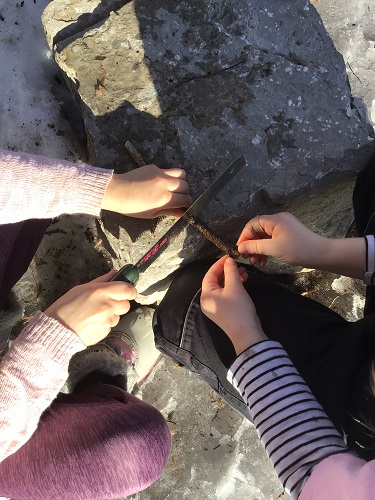 2 school age children, one holding a saw and sawing a stick, another school age child helping by holding the stick
