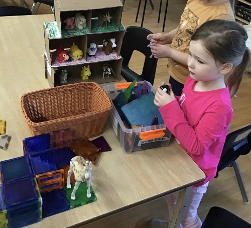 2 school age children standing at the table with a bin of magnetic tiles and animals and a small magnetic structure with animals standing on it