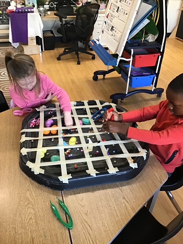 2 School age children sitting at the table with a tuff tray with tape overtop and pom poms and plastic eggs underneath, the children are holding tweezers