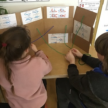 2 school age children sitting at a table with a board and different types of knots the children are attempting