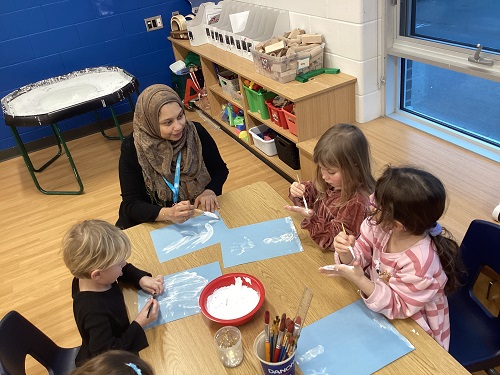 An educator and 3 school age children sitting at a table using paint to create a winter art scene