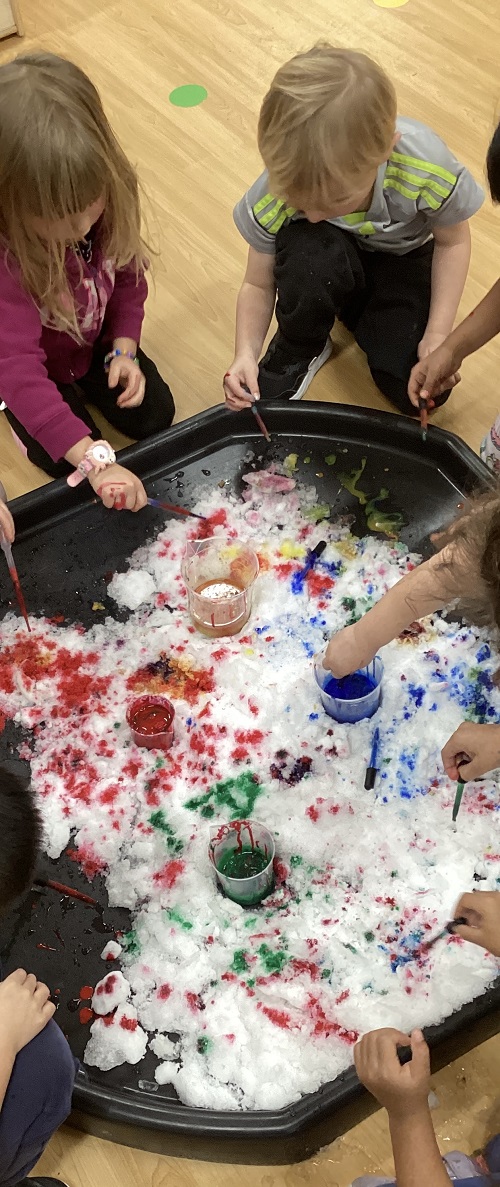 A group of school age children painting snow with various colours on a tuff tray