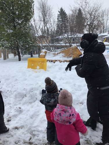 Toddler educator is throwing a snowball at the yellow sensory bin in the distance, 2 Toddler children are standing beside the educator watching the snowball being thrown