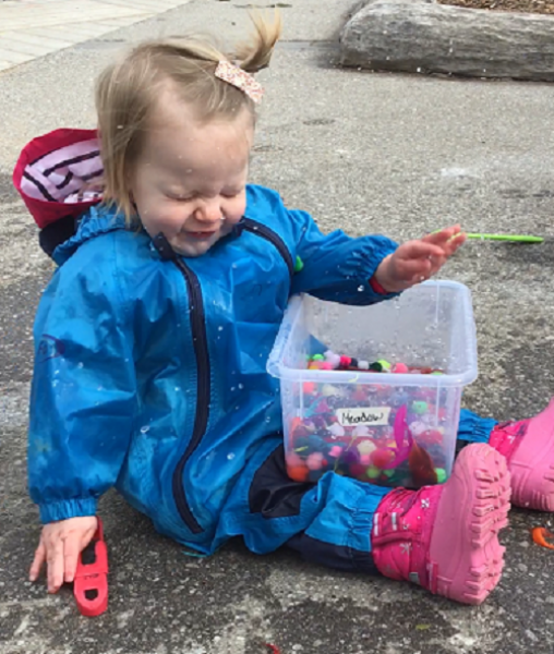 girl getting splashed sitting outside holding a bin of pom poms