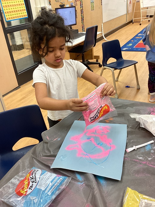 child pouring out pink sand onto a glue design on paper.