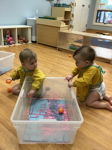 two infant girls looking into the large tub with the paint, paper and balls rolling around