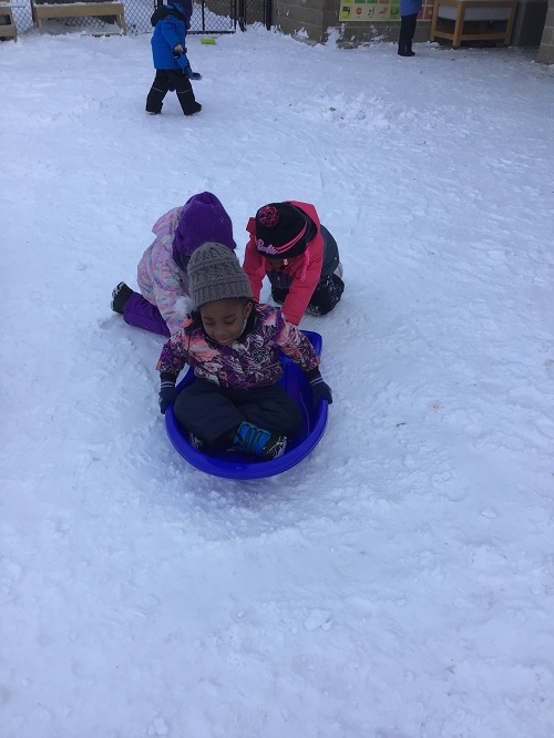 Children pushing a peer on a sled on the snowy playground.