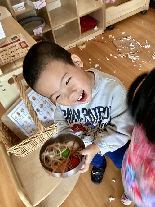A preschool child excitedly holding a bowl of play noodles and chopsticks