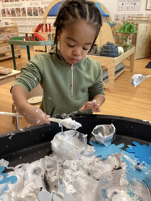 A preschool child scooping shaving cream into a small bowl