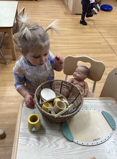A toddler in the dramatic play area with some play materials and a play baby in a chair